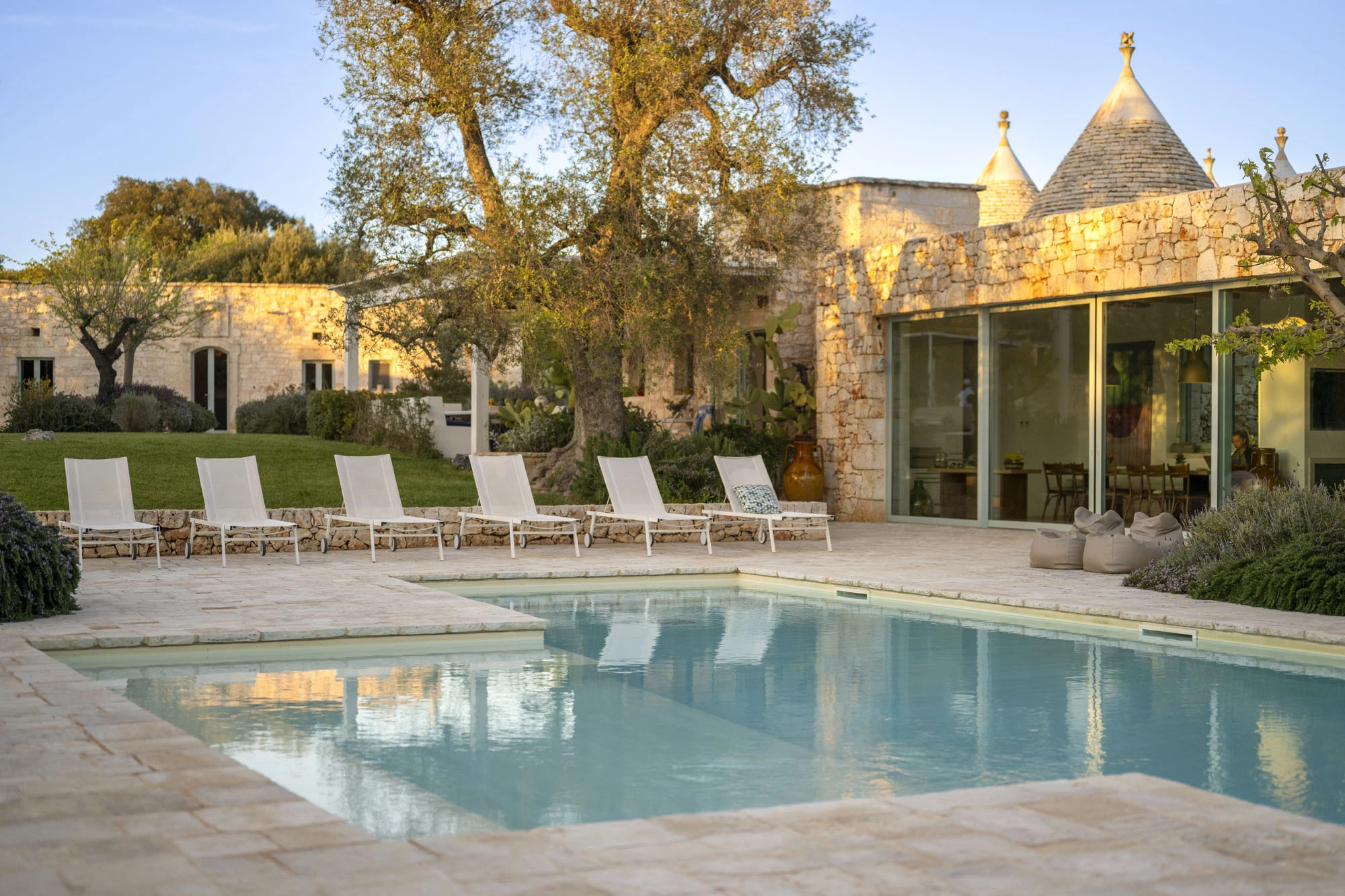 Rectangular swimming pool at golden hour with white sun loungers, glowing stone trulli villa facade, open glass living room, ancient olive trees and terracotta amphora at a luxury Puglia countryside villa