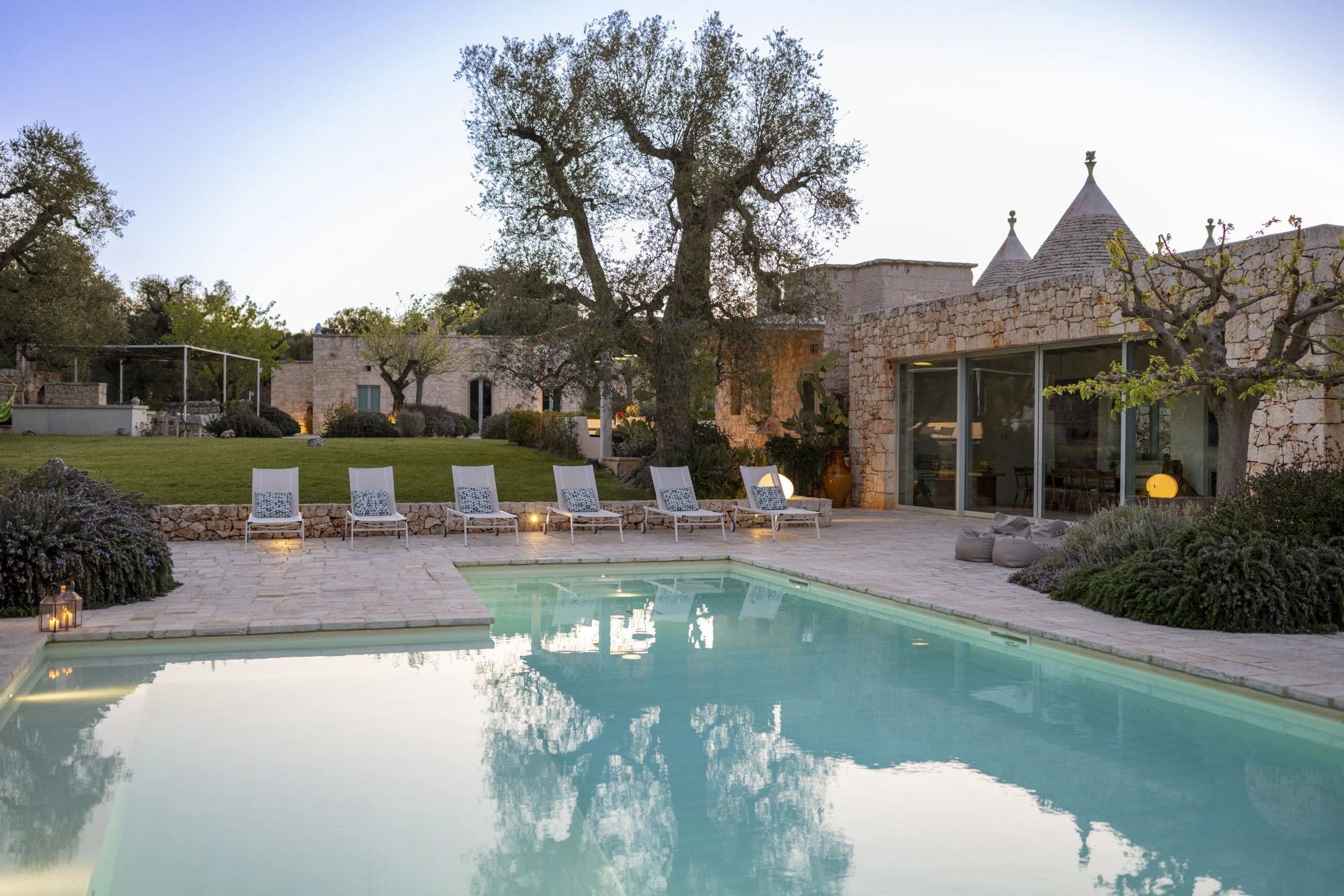 Luxury Puglia trulli villa pool at dusk with white sun loungers, garden lanterns, glowing interior living room, trulli cones, ancient olive tree and Mediterranean garden planting at twilight