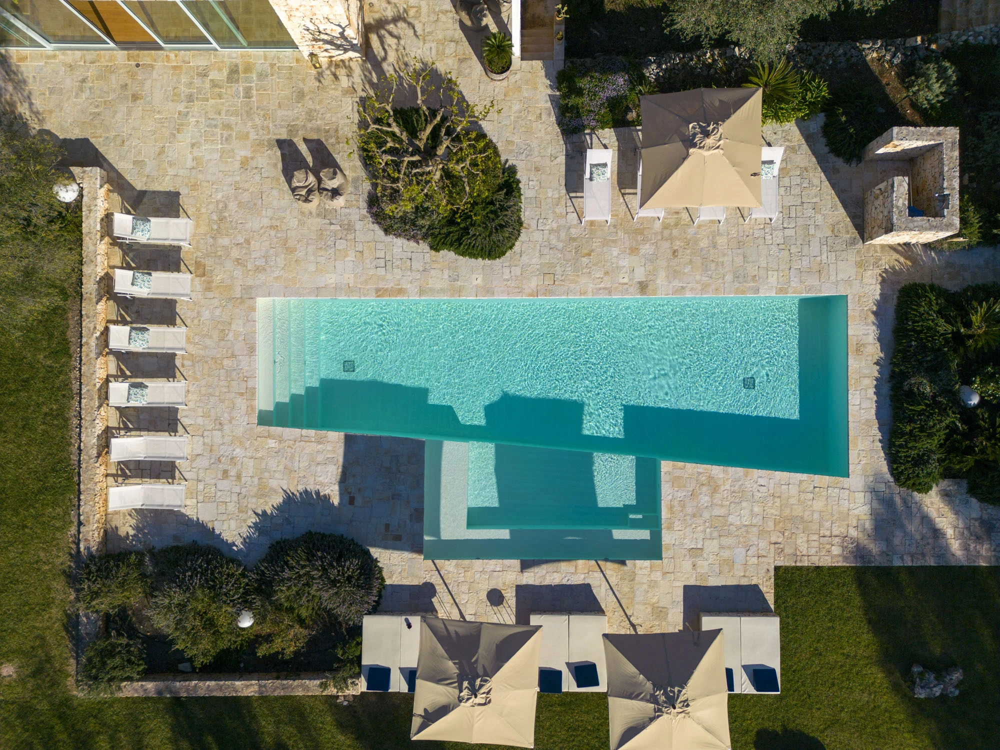 Overhead aerial drone view of a large rectangular turquoise swimming pool with multiple white sun loungers, cream parasols, limestone terrace and Mediterranean garden at a luxury Puglia villa